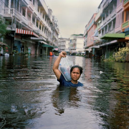 This photograph shows a young person wading through a flooded street holding a plastic bag of groceries. The water is almost up to their shoulders.