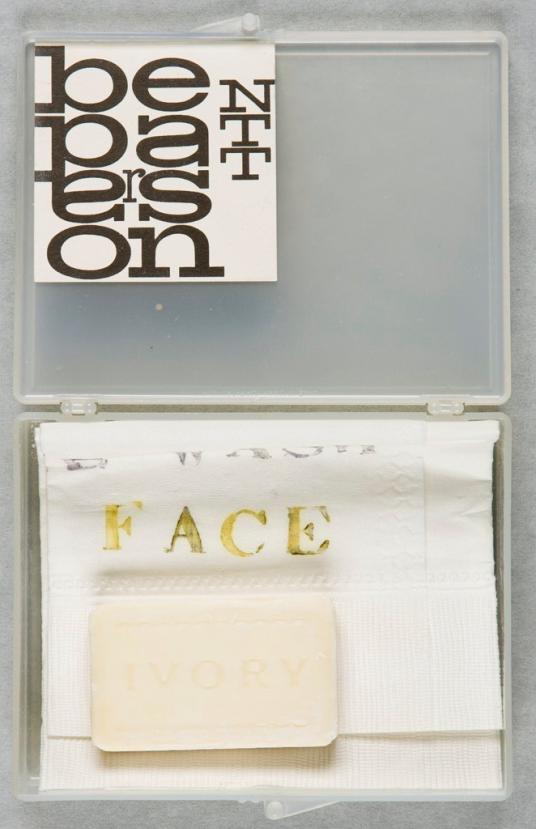 A small open translucent plastic box contains a folded paper towel and a soap bar.