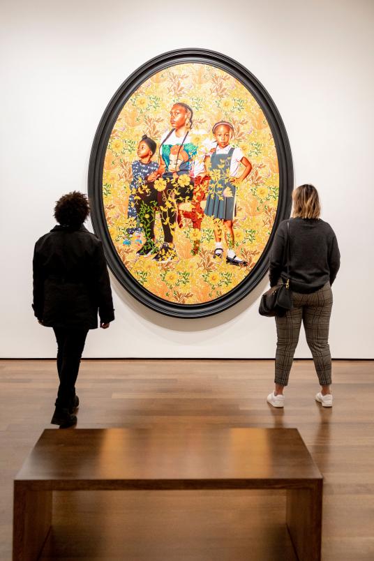 Two people stand in a gallery looking at a large oval-shaped painting of three girls surrounded by golden plants.