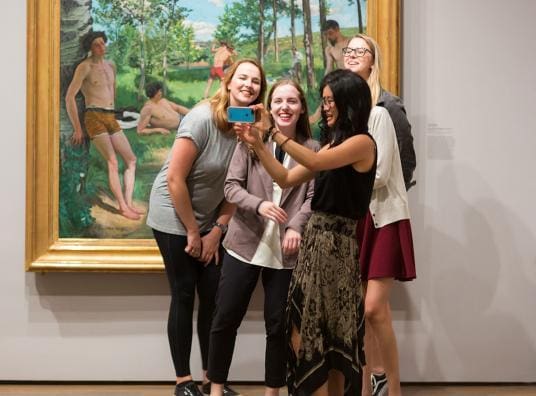 In a gallery, a group of students stand in front of a colorful landscape to take a selfie.