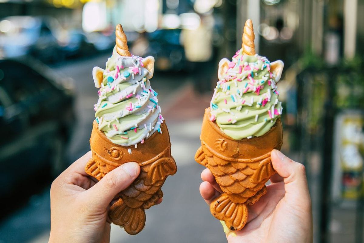 Two hands hold up two fish-shaped soft serve cones. The soft serve is pale green with rainbow sprinkles and golden unicorn horns and ears.