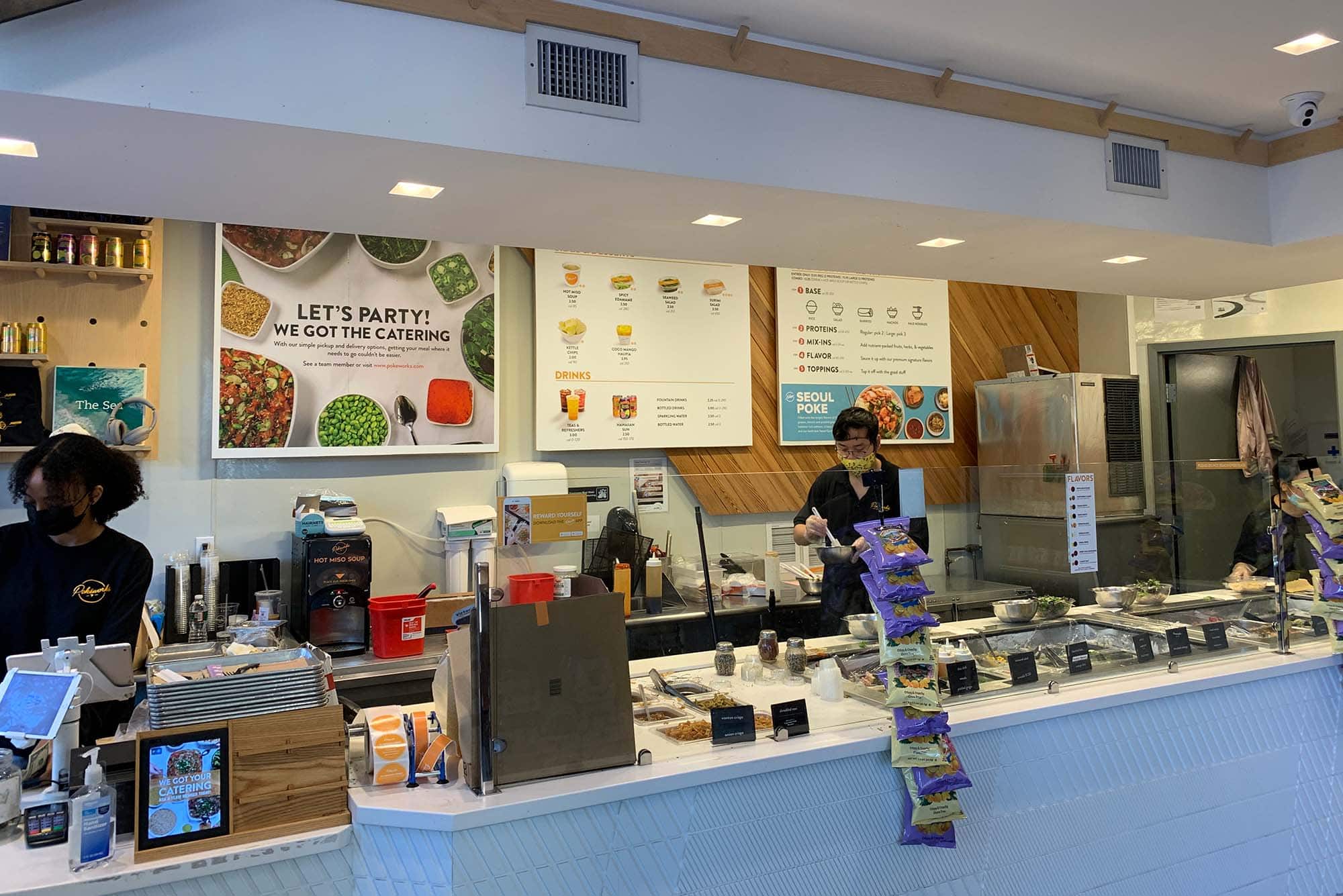 Photo taken inside Pokeworks towards the counter where people order. Two people stand behind the counter and menus are seen hanging in the background.