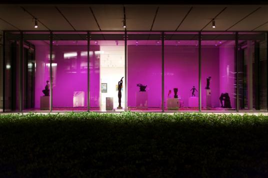 An exterior nighttime shot of a gallery illuminated by pink light, with multiple sculptures on display.
