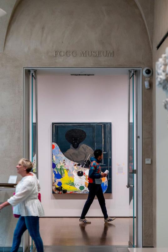 A large, colorful painting of a Black man holding a palette and paintbrush can be seen through an interior entryway to a museum gallery,. Above the entryway reads “Fogg Museum.” A person is walking in front of the painting. Outside the gallery space is a woman walking toward the left.