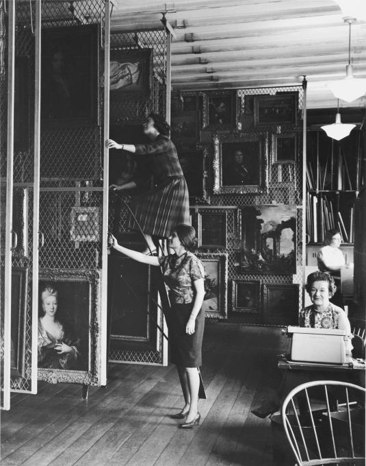 In a storage room, two students examine paintings hung on rolling racks, while two other women sit at desks.