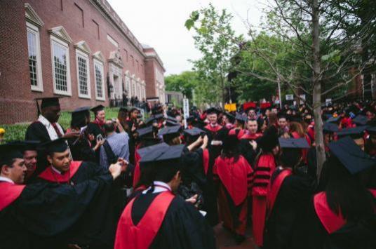 A large crowd of college graduates in black caps and gowns with red sashes gather outside near a large brick building.