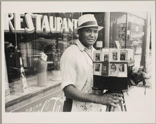 This image presents an African American photographer grasping his camera, which is covered with sample portraits, outside a restaurant.