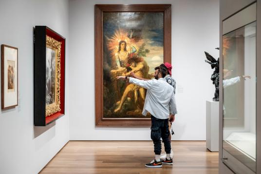 In the corner of a gallery, two people stand near a painting; one of them points toward it.