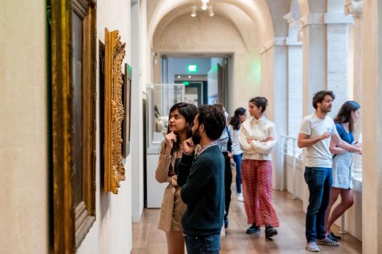 Several people in a museum arcade, some of whom are looking at works on the wall.