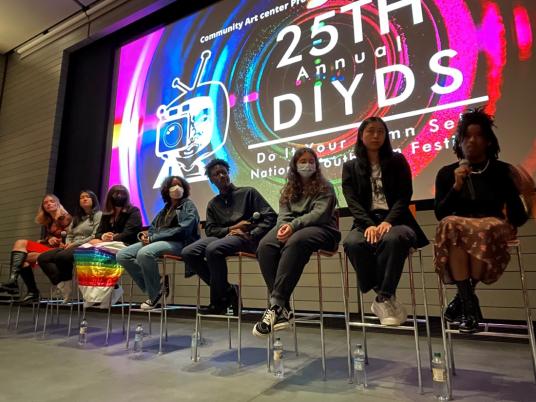 A photograph of young people on stage in front of a large screen that reads “25th Annual DIYDS.”