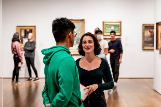 Two people talk in a gallery. In the background art is hanging on the gallery walls.