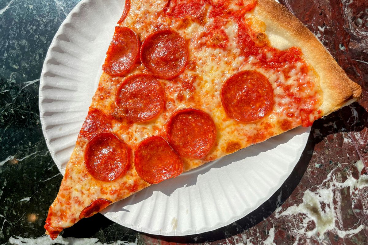 A large pepperoni and cheese slice of pizza is photographed on a white paper plate on a marble countertop.