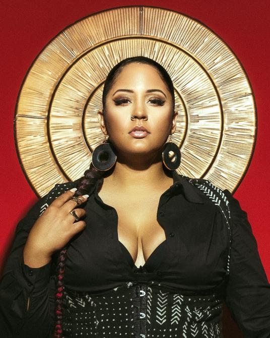 A Black woman stands in front of a deep crimson background, with a golden halo behind her head.