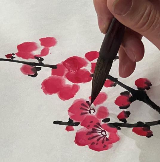 Close-up of a hand holding a brush over a painting of pink blossoms on dark twigs.