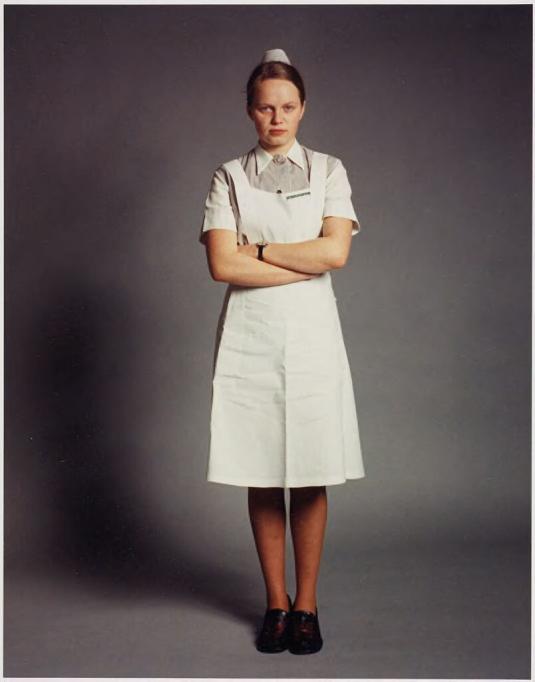 A color photograph of a person wearing a knee-length white smock and small white hat, against a neutral gray background.