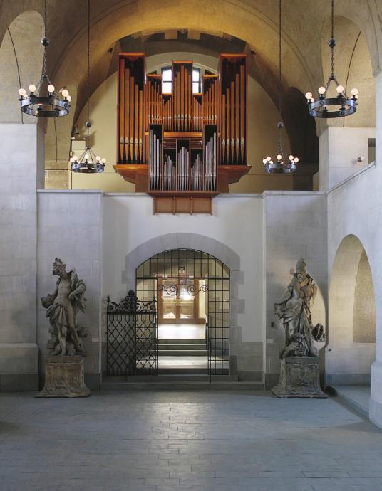 A room with several arches and figural statues in the corners with an organ musical instrument in the center.