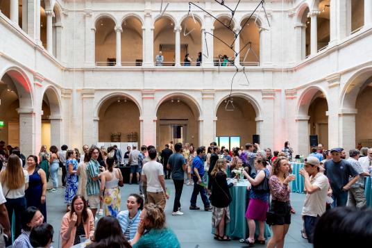 A large crowd gathers in a courtyard surrounded by arches. Small groups of people are either standing, sitting down around tables, or standing at high-top tables. A large metal sculpture hangs from the center of the courtyard.