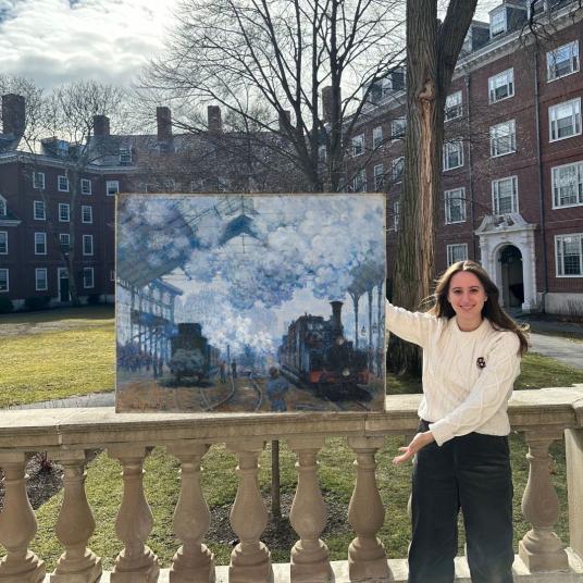 A smiling young woman stands in a campus quad gesturing toward a painting that appears to be floating on top of the balustrade behind her. The brushstrokes coalesce into a bluish scene of trains at a smoke-filled station.