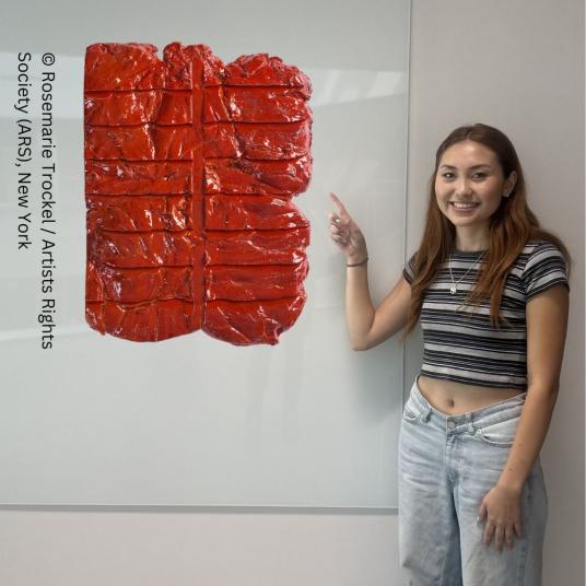 A smiling young woman stands before a whiteboard pointing toward a sculpture that appears to be floating. The sculpture is a shiny red slab scored with a line down the middle and multiple lines across.