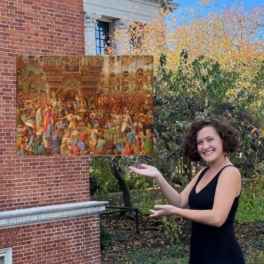 A smiling young woman stands outside a brick building gesturing toward a painting that appears to float beside her. The painting depicts, in mostly gold tones, throngs of visitors to an ornate religious structure.