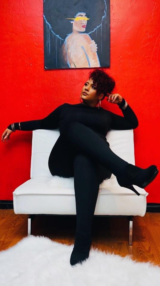 A Black woman sitting on a cushioned chair underneath a painting on a red wall.