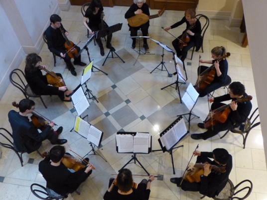 Eleven musicians dressed in black play stringed instruments in a circle.