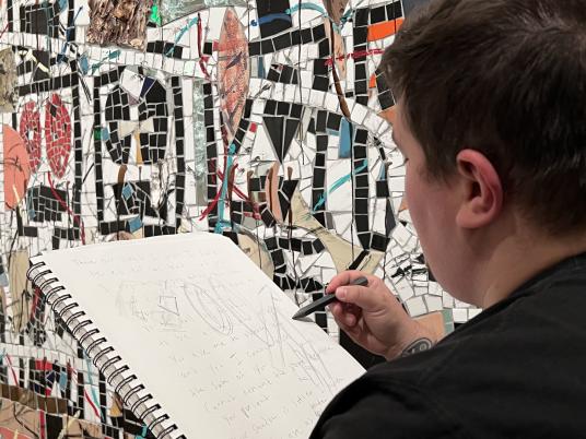A person sketching in front of a colorful wall with mosaics.