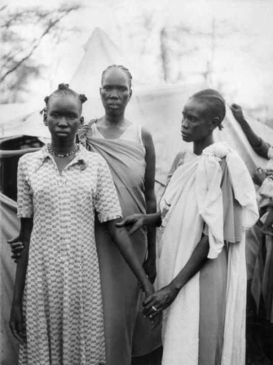 A black and white photograph of three people outdoors, wearing long dresses. One of the people touches the arm of another with both hands.