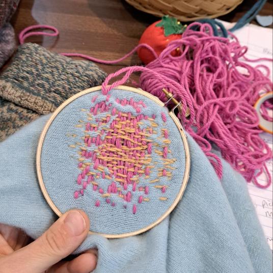 A hand holds an embroidery hoop attached to a blue garment; inside the hoop are embroidered pink and beige threads.