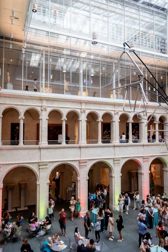 A bird’s-eye view of groups of people gathering in a courtyard surrounded by arches. Art is visible on the walls on the second and third floors.
