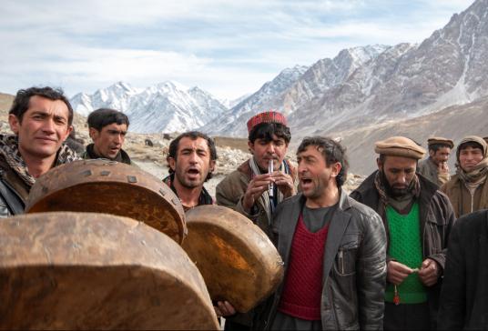 In a colorful photograph, several men play music or sing, with snow-covered mountains behind them.