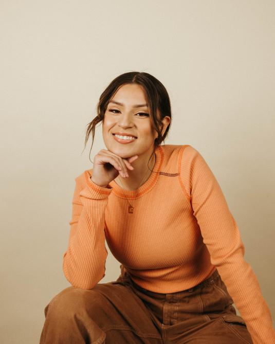A smiling young woman in an orange shirt and brown pants sits on a stool.