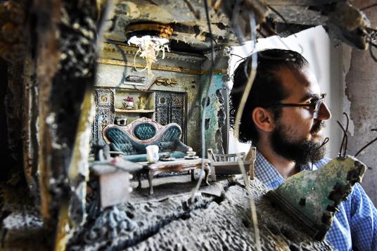 A bearded man stands next to a representation in miniature of the remains of a living room in a war-ravaged building.