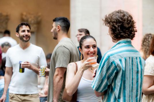 Two people converse in a courtyard setting. The woman facing the camera holds a drink. Two men are chatting in the background.