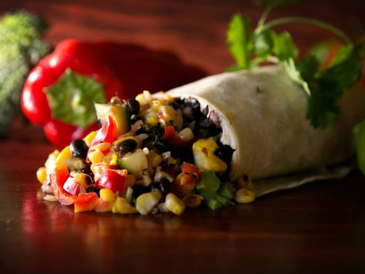 Corn, black beans, and red pepper pieces spill out of burrito on a dark wood surface. A red pepper and cilantro sit in the background.
