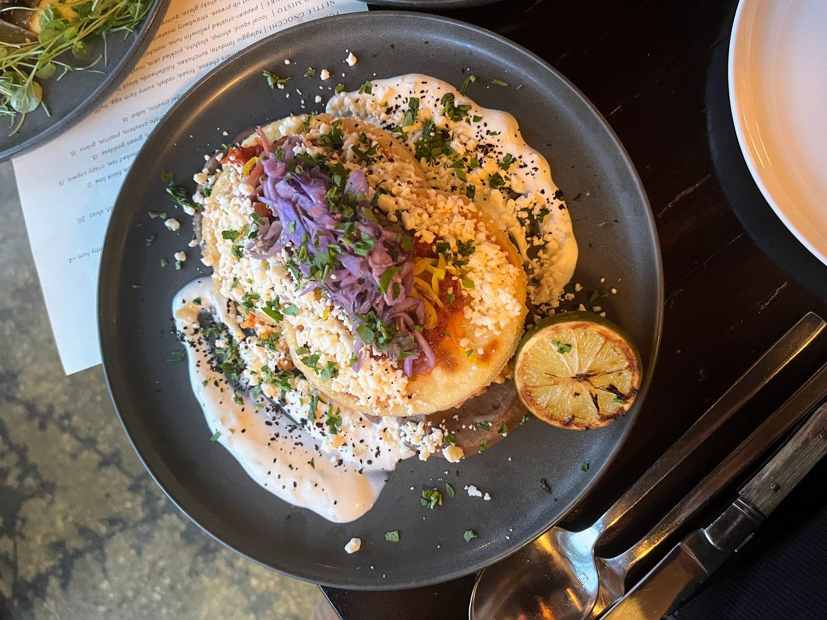 Two arepas in a white sauce with pickled vegetables and cheese sprinkled over top.