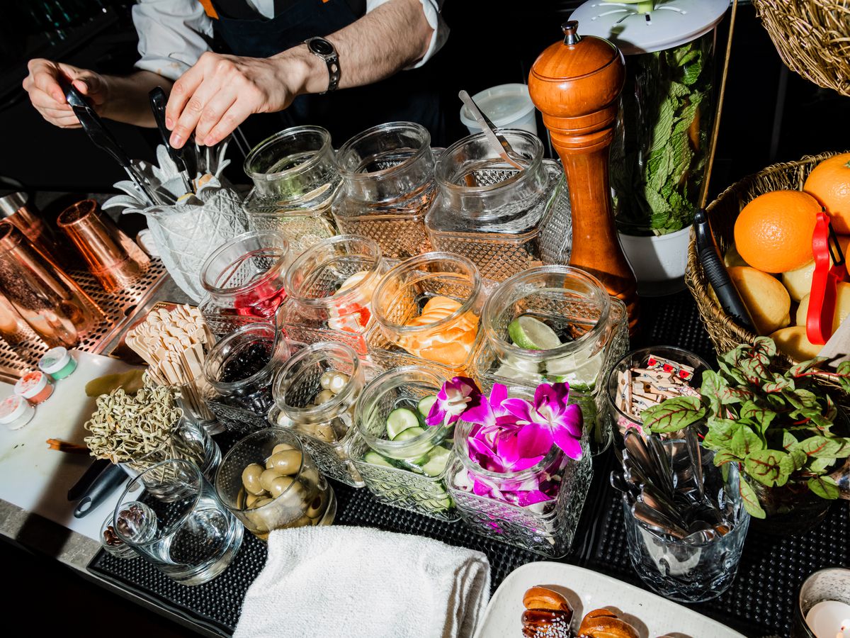Two people stand on either sides of a bar, both in reaching distance of a bunch of containers holding colorful fruits, sticks, glitter, and a variety of other drink toppings.