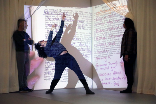 A young woman strikes a dance pose; book pages and words are projected onto her. 