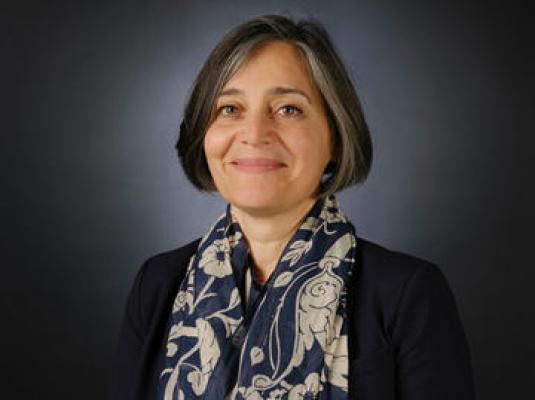 A woman with black hair smiles at the camera. She wears a black suit jacket and a dark scarf patterned with gray leaves.