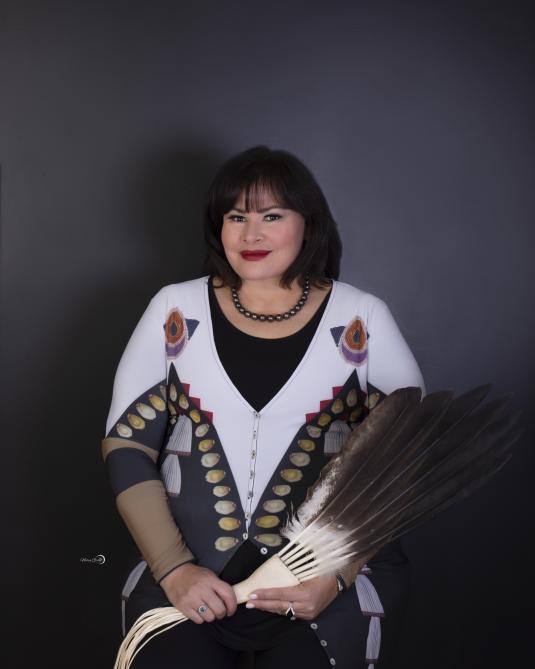 A woman with black hair is seated and holding a traditional eagle feather fan.