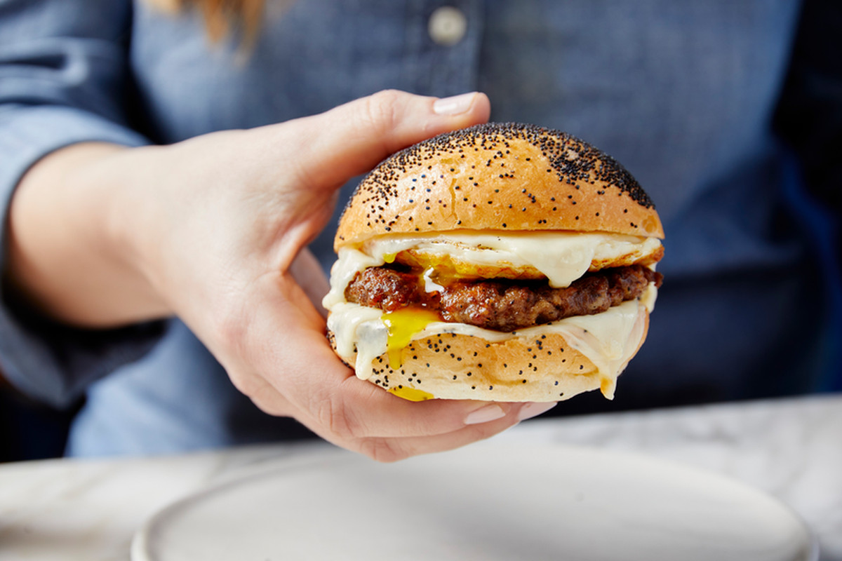 A hand holds out a cheesy breakfast sandwich on a seeded roll in front of the camera.