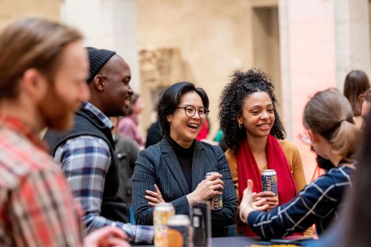 A group of people are gathered around a high table, holding drinks and smiling. The background is softly blurred.