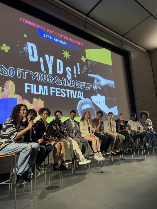 A group of teens is seated on a panel, and one teen in a striped shirt is speaking into a microphone. A screen behind them reads “DIYDS!! Do It Your Damn Self!! Film Festival.”